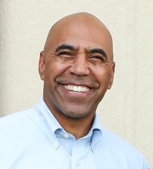 Smiling professional man in a light blue button-down shirt standing against a beige background.