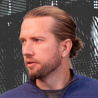 Professional man with beard and pulled-back hair wearing a blue sweater, looking to the side in front of a modern background.